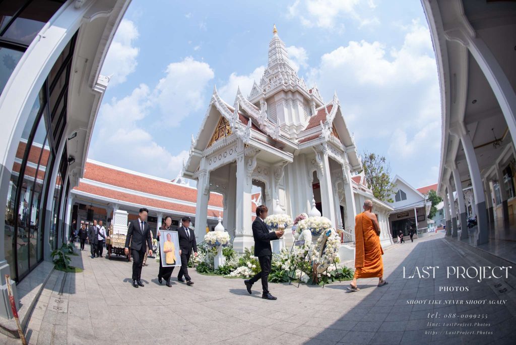 พิธีเวียนศพรอบเมรุในงานฌาปนกิจศพแบบไทย นำโดยพระสงฆ์ ญาติถือรูปภาพและกระถางธูป พร้อมขบวนหีบศพหน้าเมรุวัด A Buddhist funeral procession circling the white crematorium in a Thai temple, led by a monk followed by family members carrying a portrait, incense, and the casket