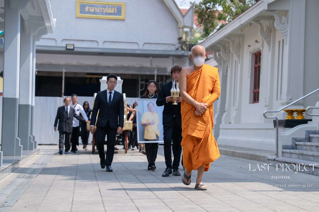 พระสงฆ์เดินนำขบวนเวียนเมรุในพิธีฌาปนกิจศพ โดยมีครอบครัวถือรูปผู้วายชนม์และเคลื่อนหีบศพภายในวัดไทย A Buddhist monk leading a funeral procession with family members holding a portrait of the deceased and moving the casket around the crematorium at a Thai temple