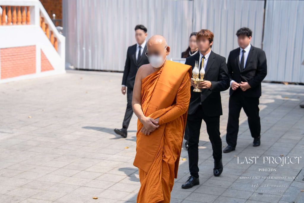 พระสงฆ์สวมหน้ากากอนามัยเดินนำขบวนเวียนรอบเมรุในพิธีฌาปนกิจศพ ณ วัดไทย โดยมีญาติและผู้เข้าร่วมพิธีในชุดสูทสีดำเดินตามหลังท่ามกลางแสงแดดธรรมชาติภายนอกอาคาร A Buddhist monk wearing a face mask leads a funeral procession walking around a crematorium at a Thai temple, followed by mourners dressed in black suits, captured outdoors during a traditional Thai cremation ceremony.