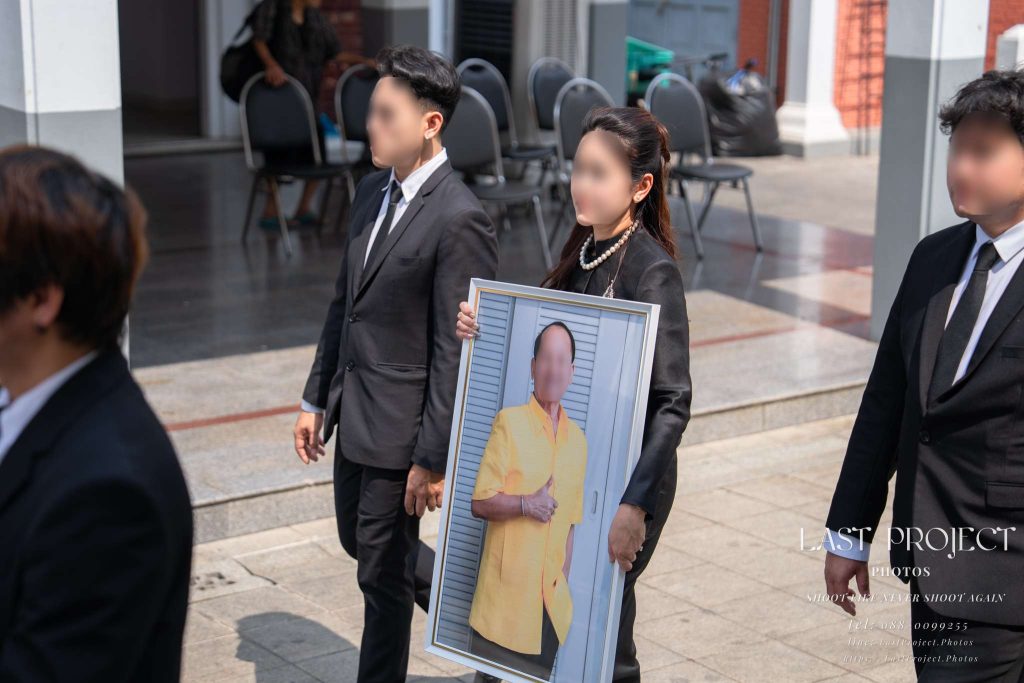 ภาพหญิงและชายไทยในชุดดำไว้ทุกข์ร่วมเดินขบวนแห่ในพิธีฌาปนกิจศพที่วัดไทย โดยผู้หญิงถือภาพถ่ายของผู้ล่วงลับอย่างสงบเสงี่ยมและเคารพรัก Thai mourners in black attire walking in a funeral procession at a temple, with a woman respectfully carrying a framed portrait of the deceased.