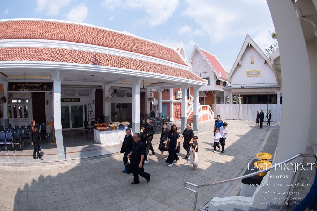 ภาพถ่ายมุมกว้างเลนส์ฟิชอายในงานพิธีฌาปนกิจศพ ขณะแขกสวมชุดดำกำลังเดินในลานวัดไทย Wide-angle fisheye perspective of guests in black attire walking in a Thai temple courtyard during a Buddhist cremation ceremony