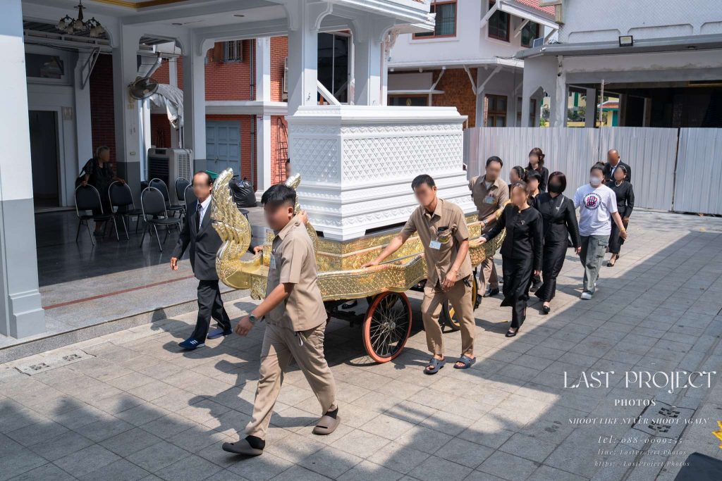 ภาพพิธีเวียนศพรอบเมรุในวัดไทย เจ้าหน้าที่และแขกผู้ร่วมงานแต่งกายชุดดำร่วมกันเข็นราชรถแห่หีบศพสีขาวทองในงานฌาปนกิจศพ A traditional Thai Buddhist cremation ceremony at a temple, where temple staff and mourners in black attire collectively push an ornate white and gold funeral carriage carrying a casket during the circumambulation around the crematorium.