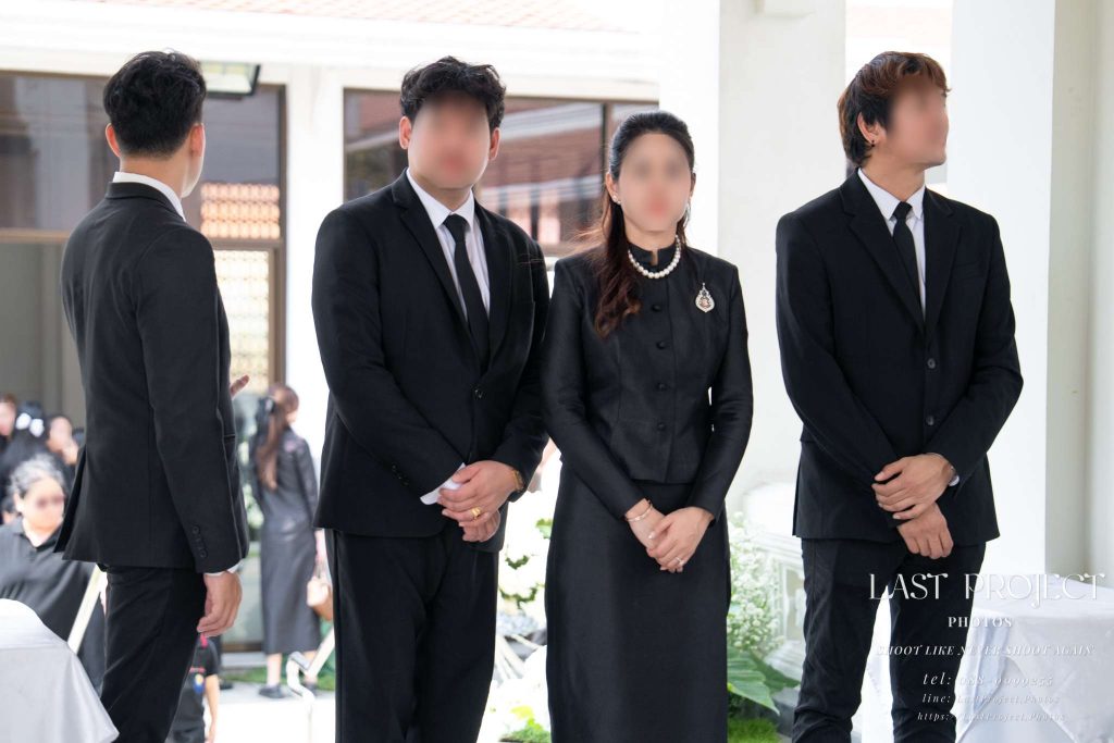 ภาพถ่ายเจ้าภาพและครอบครัวในชุดไว้ทุกข์สีดำยืนรอประกอบพิธีฌาปนกิจบนเมรุวัดไทย A Thai Buddhist funeral host family in black mourning attire standing at the temple crematorium awaiting the casket during a cremation ceremony
