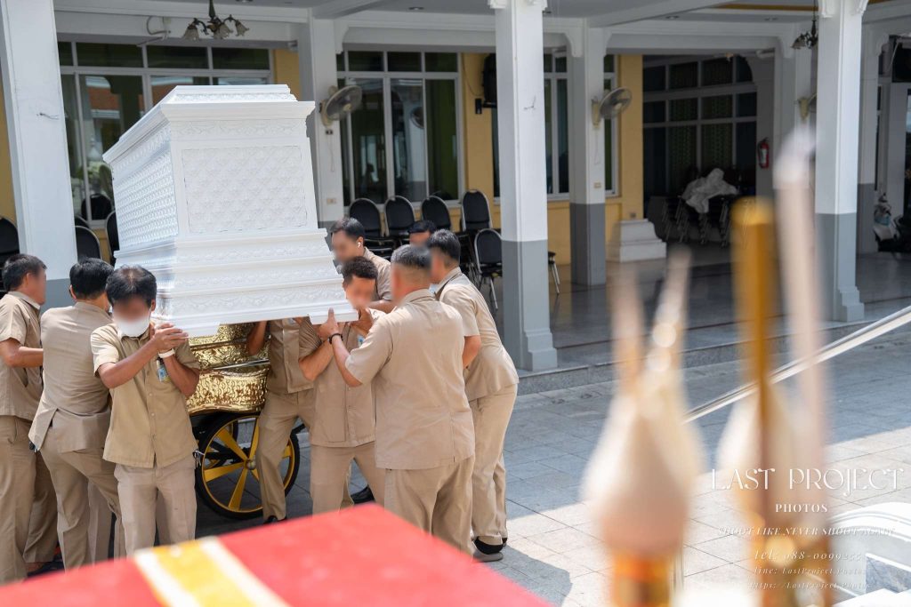 เจ้าหน้าที่กำลังยกหีบศพสีขาวขึ้นสู่เมรุในพิธีฌาปนกิจ ณ วัดไทย Funeral attendants lifting a white casket onto the crematorium during a Thai Buddhist cremation ceremony