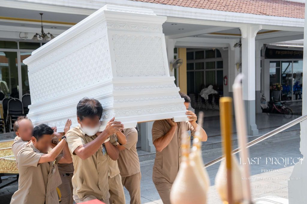เจ้าหน้าที่วัดกำลังแบกหีบศพสีขาวขึ้นเมรุในพิธีฌาปนกิจ ณ วัดไทย Temple staff carrying a traditional white casket to the crematorium for a Thai Buddhist funeral ceremony