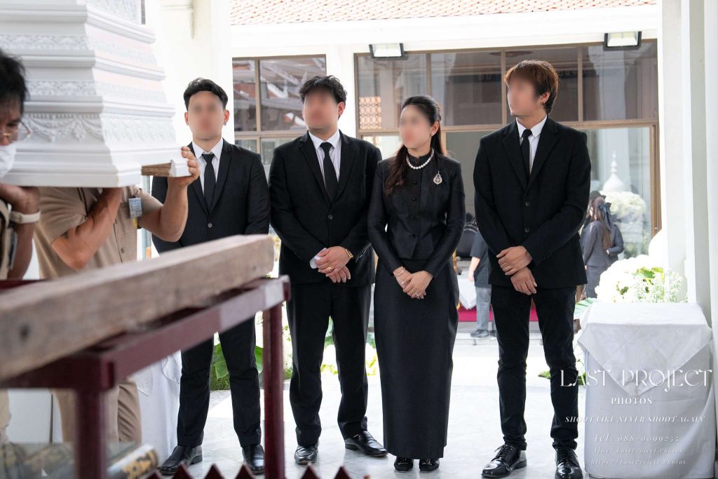 เจ้าภาพในชุดไว้ทุกข์สีดำยืนรอรับหีบศพในพิธีฌาปนกิจศพ ณ เมรุวัดไทย Host family in black mourning attire awaiting the casket at a Thai Buddhist temple crematorium