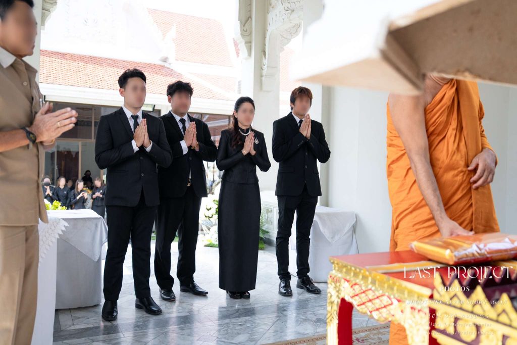 เจ้าภาพและครอบครัวในชุดดำพนมมือสงบนิ่งขณะพระสงฆ์ประกอบพิธีทอดผ้าไตรในงานฌาปนกิจศพ ณ วัดไทย Host family dressed in black standing with hands clasped in prayer while a Buddhist monk receives funeral robes during a Thai Buddhist cremation ceremony at a temple
