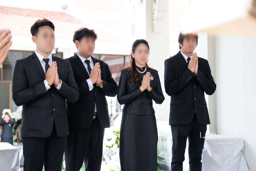 ภาพเจ้าภาพและญาติสวมชุดดำยืนพนมมือในพิธีฌาปนกิจศพวัดไทย Thai hosts and family members in black suits standing with hands clasped in prayer (Wai) during a Buddhist funeral and cremation ceremony at a Thai temple