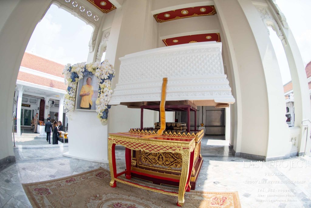 ภาพถ่ายมุมกว้างหีบศพสีขาวและแท่นพิธีในงานฌาปนกิจ ณ เมรุวัดไทย A wide-angle perspective of an ornate white casket placed on a golden ceremonial platform during a traditional Thai Buddhist cremation ceremony at a temple crematory.