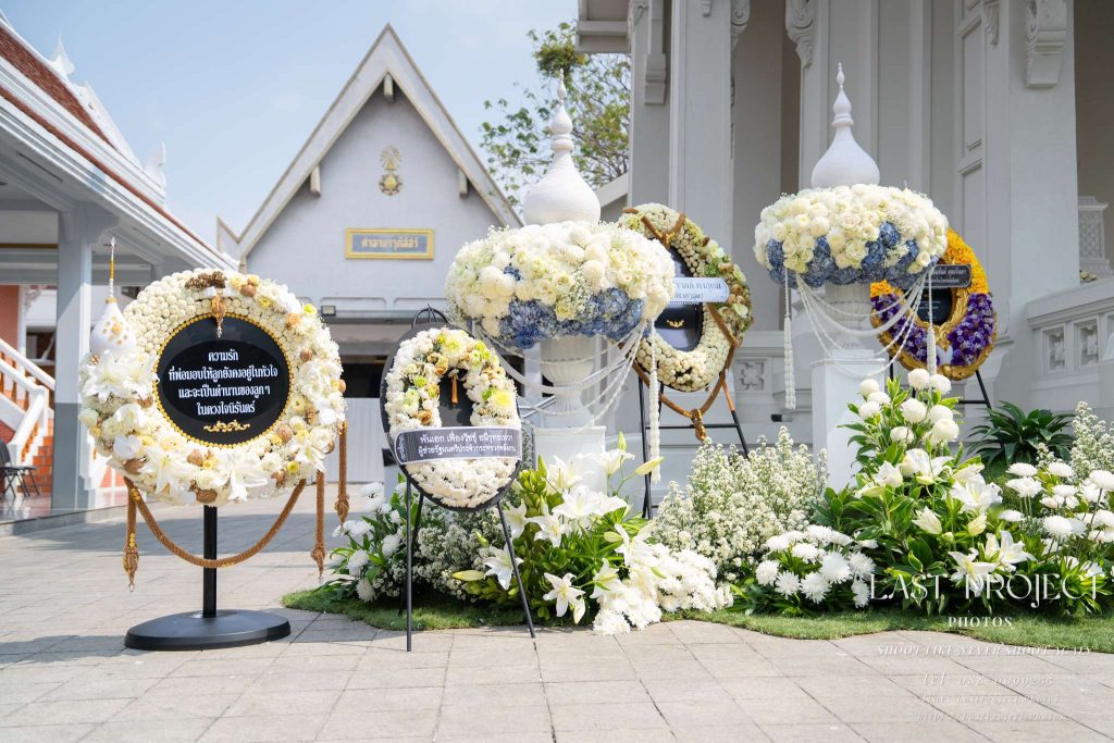 พวงหรีดดอกไม้สดสีขาวและสีฟ้าตกแต่งหน้าเมรุวัดไทยในพิธีพระราชทานเพลิงศพ Fresh white and blue floral funeral wreaths and standing flower arrangements in front of a Thai temple crematorium during a Buddhist cremation ceremony