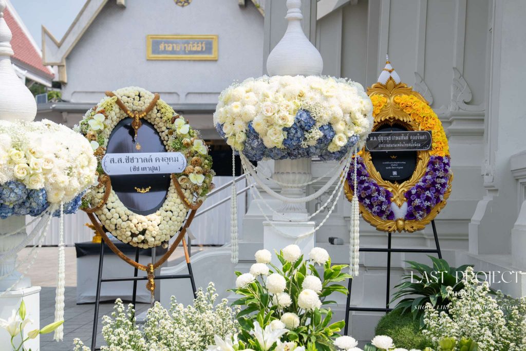 ภาพพวงหรีดดอกไม้สดหลากสีสันตั้งประดับหน้าเมรุวัดไทยในงานฌาปนกิจศพ มีป้ายชื่อผู้ส่งชัดเจนเพื่อการบันทึกภาพของเจ้าภาพ Colorful funeral wreaths with donor name banners displayed in front of a Thai temple crematorium during a cremation ceremony, showcasing traditional Thai mourning flowers for remembrance.