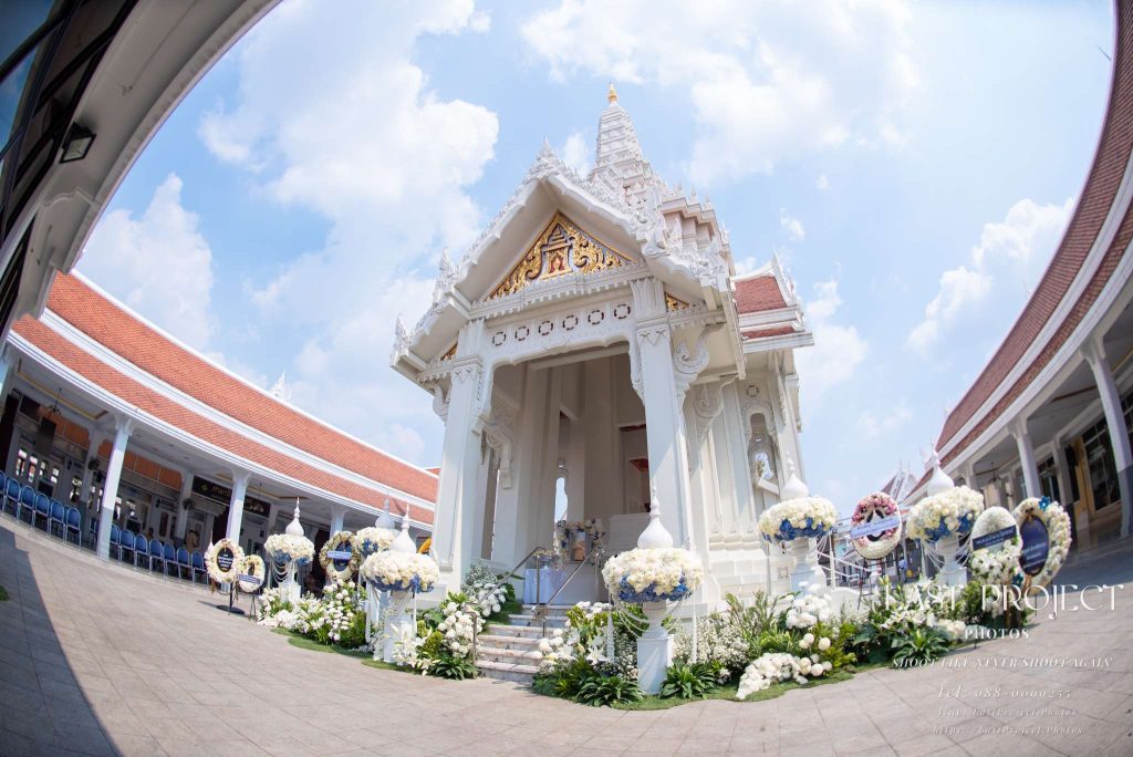ภาพถ่ายมุมกว้างพิเศษเมรุวัดไทยสีขาวประดับตกแต่งด้วยดอกไม้งานศพและพวงหรีดอย่างสมเกียรติในพิธีฌาปนกิจ Ultra wide-angle view of a white Thai temple crematorium decorated with funeral flower arrangements and wreaths for a Buddhist cremation ceremony