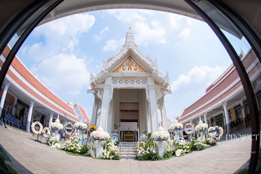 ภาพถ่ายเมรุวัดไทยในพิธีศพด้วยเลนส์มุมกว้างพิเศษ (Fisheye) ตกแต่งด้วยดอกไม้สีขาวรอบบันไดทางขึ้นและพวงหรีด มีศาลาสองข้างทางและท้องฟ้าแจ่มใส A fisheye wide-angle photograph of a traditional Thai temple crematorium (Meru) decorated with white flowers and wreaths for a funeral ceremony, framed by temple pavilions on both sides under a bright blue sky.