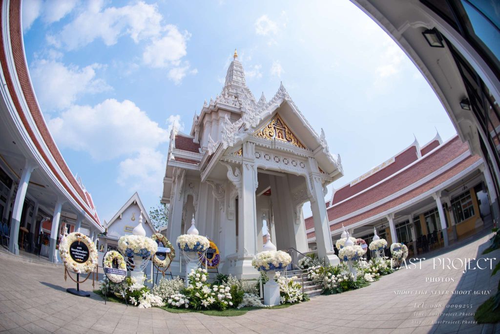 ภาพมุมกว้างเลนส์ฟิชอายเมรุวัดไทยตกแต่งดอกไม้และพวงหรีดในพิธีฌาปนกิจวันฟ้าโปร่ง A wide-angle fisheye shot of a white Thai temple crematorium decorated with funeral floral arrangements and wreaths on a clear day.