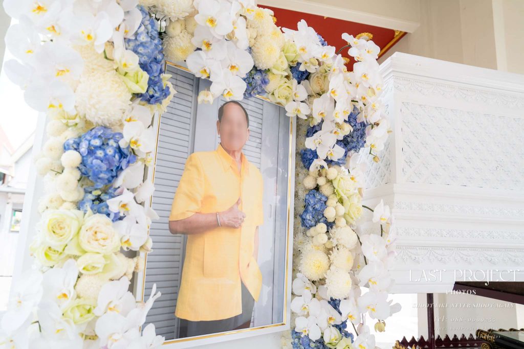 กรอบรูปหน้าศพประดับซุ้มดอกไม้สีขาวและสีฟ้าข้างหีบศพสีขาวในพิธีฌาปนกิจศพแบบไทย Portrait of the deceased adorned with white and blue funeral flower decoration placed beside the white casket in a traditional Thai Buddhist cremation ceremony