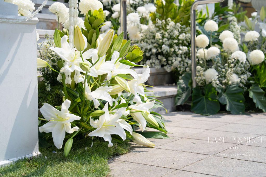 ภาพถ่ายเจาะดอกไม้สดสีขาวประดับตกแต่งบันไดทางขึ้นเมรุในพิธีฌาปนกิจ ณ วัดไทย Close-up photograph of fresh white floral arrangements decorating the crematorium steps at a Thai Buddhist funeral ceremony