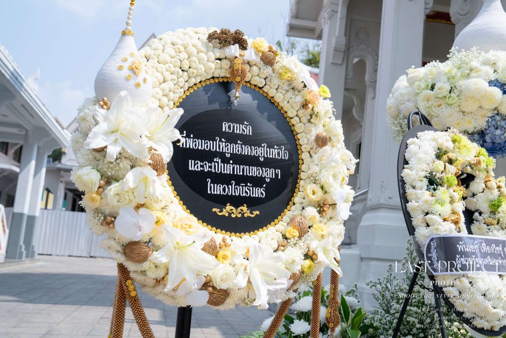 พวงหรีดดอกไม้สดโทนสีขาวทองประดับงานฌาปนกิจ ณ วัดไทย มุมมองเสยแสดงความสง่างาม Elegant white and gold fresh flower funeral wreath for a Thai cremation ceremony at a Buddhist temple captured from a low angle