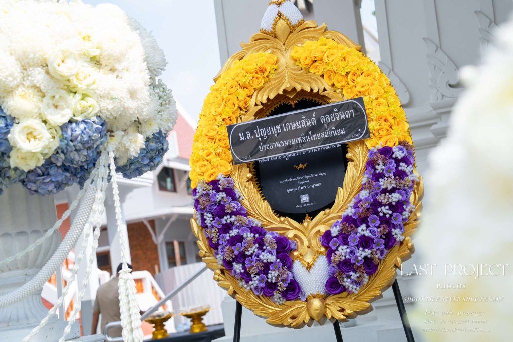พวงหรีดดอกไม้ประดับกรอบลายไทยสีทองในพิธีฌาปนกิจ ณ วัดไทย ถ่ายเจาะมุมมองผ่านโฟร์กราวด์ดอกไม้หน้าเมรุ Ornate Thai funeral wreath with gold carved frame and vibrant flowers at a temple cremation ceremony framed by foreground flowers