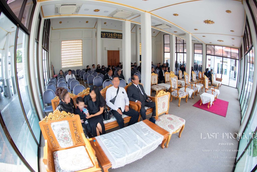 บรรยากาศภายในศาลาบำเพ็ญกุศล วัดเทพศิรินทราวาส กรุงเทพมหานคร มีแขกผู้มีเกียรตินั่งรอเข้าร่วมพิธีฌาปนกิจบนเก้าอี้และโซฟาไม้แกะสลัก ถ่ายด้วยเลนส์มุมกว้าง Interior of the pavilion at Wat Debsirindrawas, Bangkok, with guests seated on chairs and carved wooden sofas waiting for the Thai cremation ceremony, captured in a wide-angle shot.
