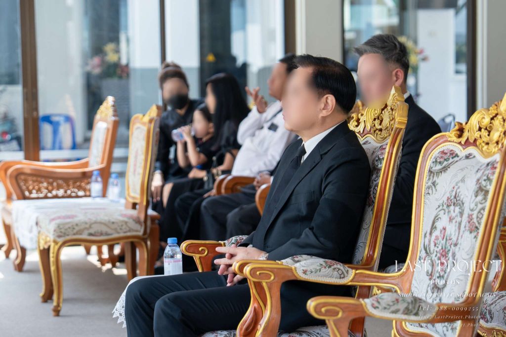 แขกผู้มีเกียรติสวมชุดดำไว้ทุกข์นั่งพับมือรอบนเก้าอี้หลุยส์ในศาลาบำเพ็ญกุศลติดแอร์ วัดเทพศิรินทร์ กรุงเทพฯ เพื่อรอพิธีฌาปนกิจ Guests in black mourning attire sitting on ornate chairs inside the air-conditioned pavilion, waiting for a Thai cremation ceremony at Wat Debsirin, Bangkok.