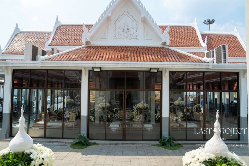ภาพบรรยากาศภายนอกศาลาพิธีธรรมในงานฌาปนกิจ ณ วัดเทพศิรินทราวาส กรุงเทพมหานคร ถ่ายด้วยเลนส์ระยะปกติ เผยให้เห็นสถาปัตยกรรมไทยอันงดงามและดอกไม้ไว้อาลัยผ่านกระจก Exterior view of a funeral pavilion during a cremation ceremony at Wat Debsirindrawas in Bangkok, captured with a normal lens, showing the ornate Thai architecture and memorial floral tributes through the glass facade.