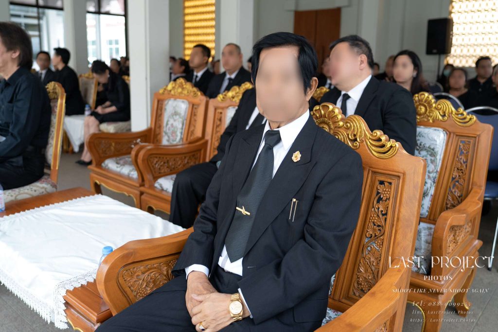 ภาพถ่ายแขกผู้มีเกียรติสวมชุดสูทสากลสีดำไว้อาลัยนั่งบนเก้าอี้ไม้แกะสลักในศาลาบำเพ็ญกุศล พิธีฌาปนกิจ วัดเทพศิรินทร์ กรุงเทพมหานคร Distinguished guest in a black formal suit seated on an ornate carved wooden chair inside the funeral pavilion during a Thai Buddhist cremation ceremony at Wat Thepsirin Bangkok