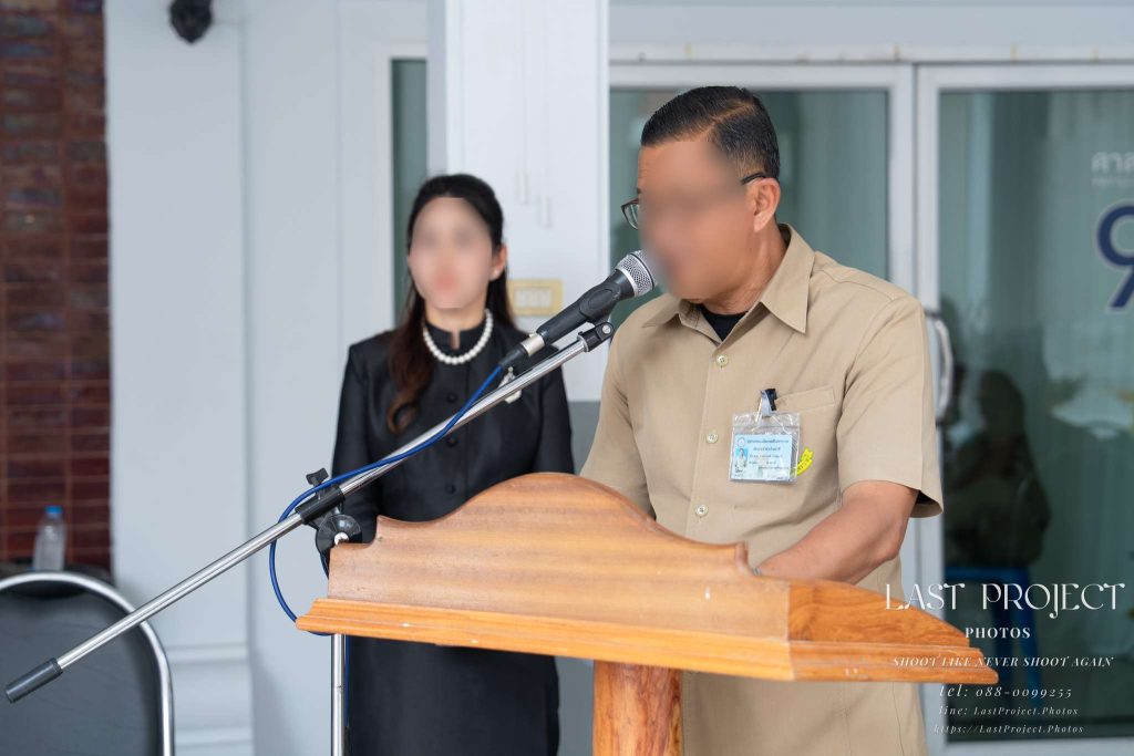 พิธีกรชายกำลังกล่าวเปิดงานบนโพเดียมไมโครโฟนในพิธีฌาปนกิจศพ ณ วัดไทย The master of ceremonies speaking into a microphone at the podium during a Buddhist funeral cremation ceremony at a Thai temple.