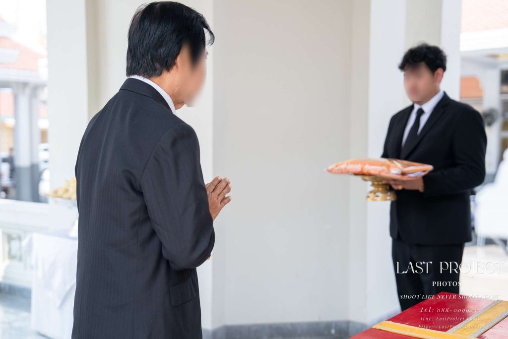 แขกผู้มีเกียรติยกมือไหว้เคารพศพบนเมรุและเตรียมรับผ้าไตรบังสุกุลจากเจ้าภาพในพิธีฌาปนกิจ ณ วัดไทย A guest paying respects with a wai on the crematorium pavilion before receiving Bangsukon monk robes from the host at a traditional Thai funeral cremation ceremony.
