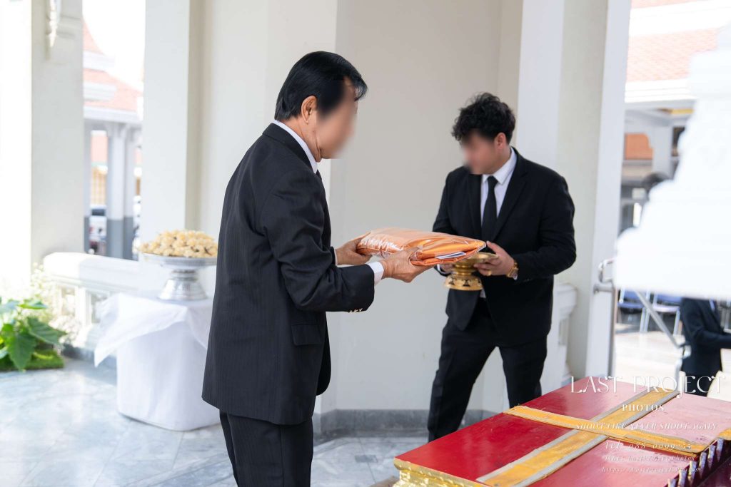 แขกรับผ้าไตรบังสุกุลจากเจ้าภาพในพิธีฌาปนกิจศพ ณ วัดไทย A guest receiving Buddhist monk robes from the host during a Thai funeral cremation ceremony at a Buddhist temple