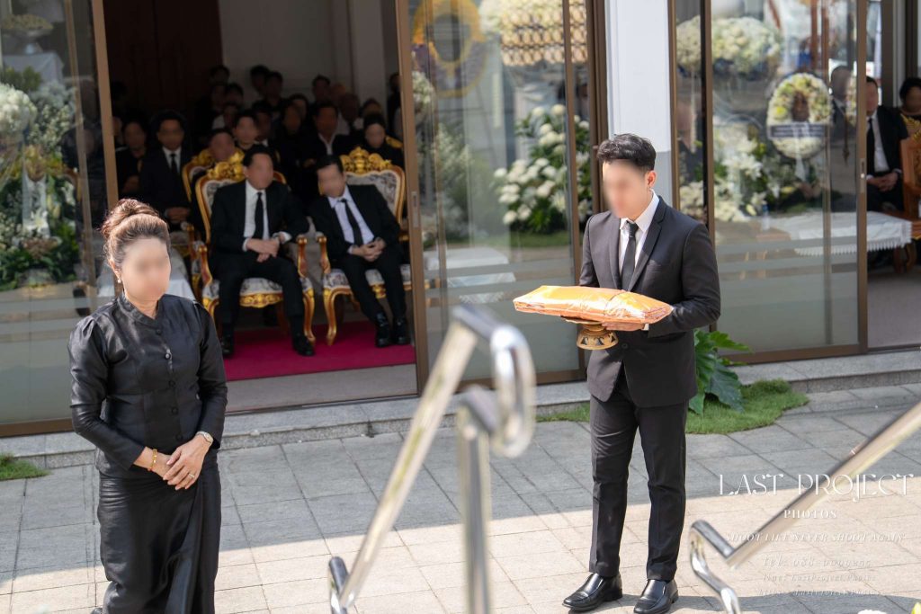 ภาพถ่ายพิธีฌาปนกิจศพขณะแขกผู้มีเกียรติถือพานผ้าไตรจีวรเดินขึ้นบันไดเมรุวัดไทย A guest in a black suit carrying yellow monk robes on a ceremonial tray up the stairs to the crematorium during a Thai funeral ceremony at a Buddhist temple.