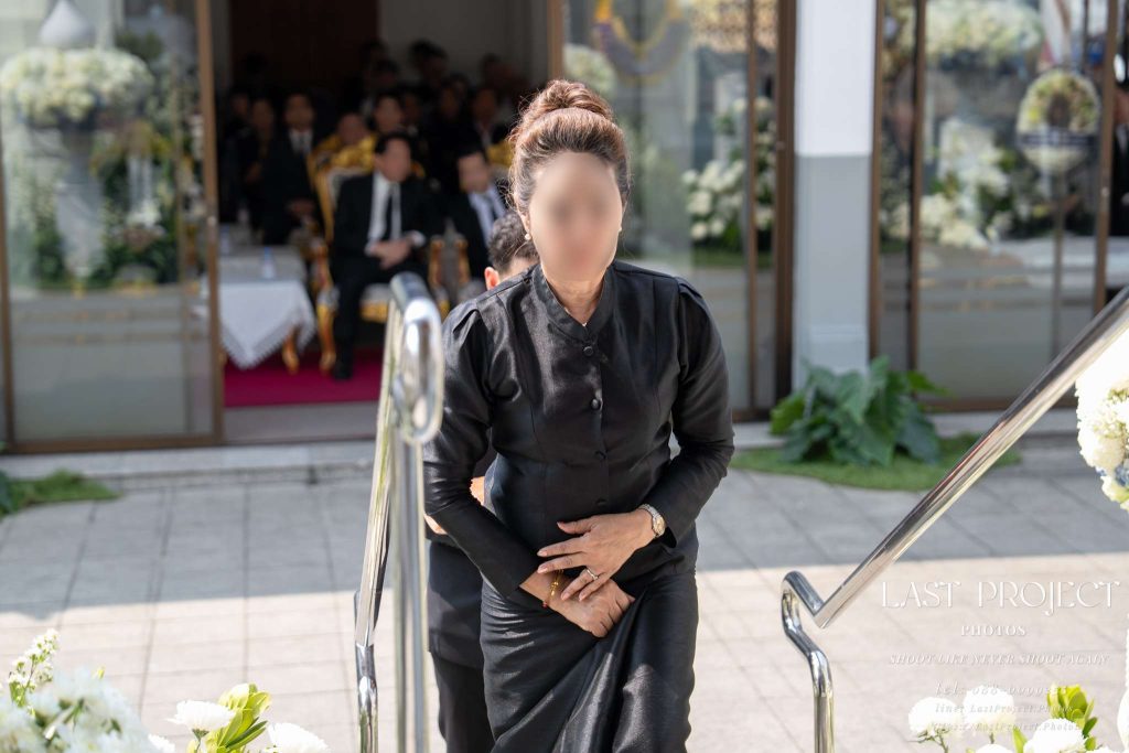 ภาพแขกสวมชุดไทยจิตรลดาไว้ทุกข์สีดำกำลังก้าวขึ้นบันไดเมรุในพิธีฌาปนกิจศพ ณ วัดไทย A guest in traditional black Thai mourning attire walking up the stairs to the crematorium during a Thai Buddhist cremation ceremony at a temple