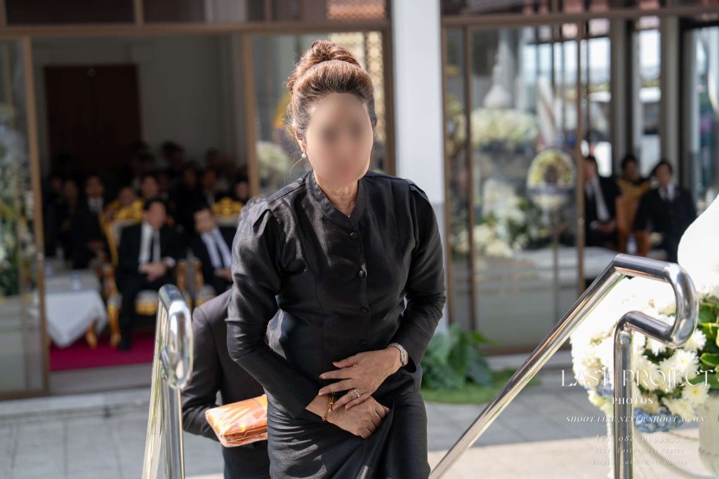 ภาพถ่ายแขกผู้มีเกียรติสวมชุดดำกำลังเดินขึ้นบันไดเมรุเพื่อประกอบพิธีทอดผ้าบังสุกุลในงานฌาปนกิจ ณ วัดไทย Photograph of an honored guest in black mourning attire ascending the crematorium steps for the Bangsukula robe ceremony at a Thai temple.