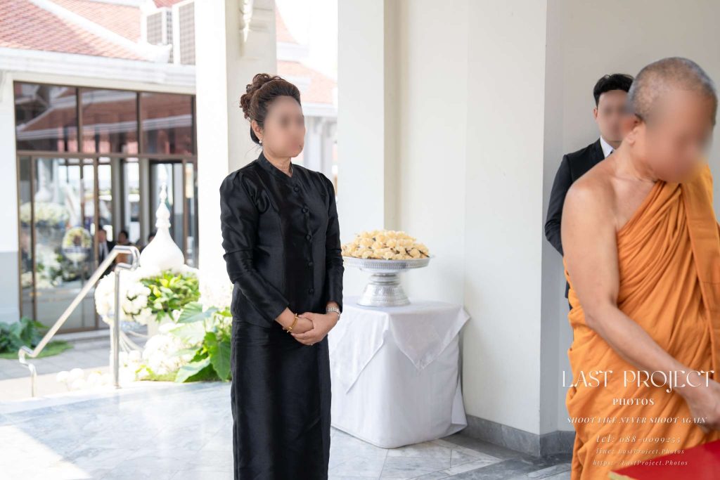 แขกผู้มีเกียรติในชุดไทยไว้ทุกข์สีดำยืนสงบนิ่งข้างพระสงฆ์ในพิธีฌาปนกิจศพ An honored guest in black Thai mourning attire standing solemnly beside a Buddhist monk during a funeral cremation ceremony