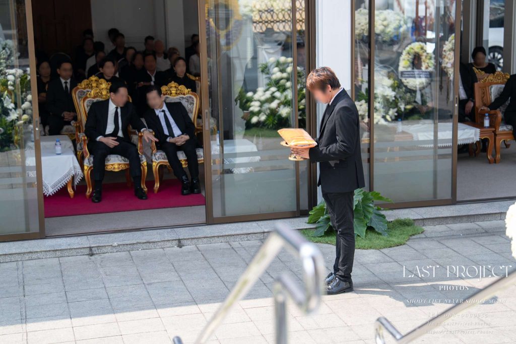 เจ้าภาพชายในชุดสูทดำยืนถือพานผ้าไตรบังสุกุลหน้าศาลาบำเพ็ญกุศลในงานฌาปนกิจศพ ณ วัดไทย มีแขกผู้ร่วมงานนั่งอยู่ด้านใน ถ่ายจากมุมสูงบันไดเมรุ A host in a black suit holding a ceremonial tray with Buddhist robes (Phas Tri) in front of a funeral pavilion during a Thai cremation ceremony at a temple, with mourners seated inside, captured from an elevated perspective near the crematorium stairs.