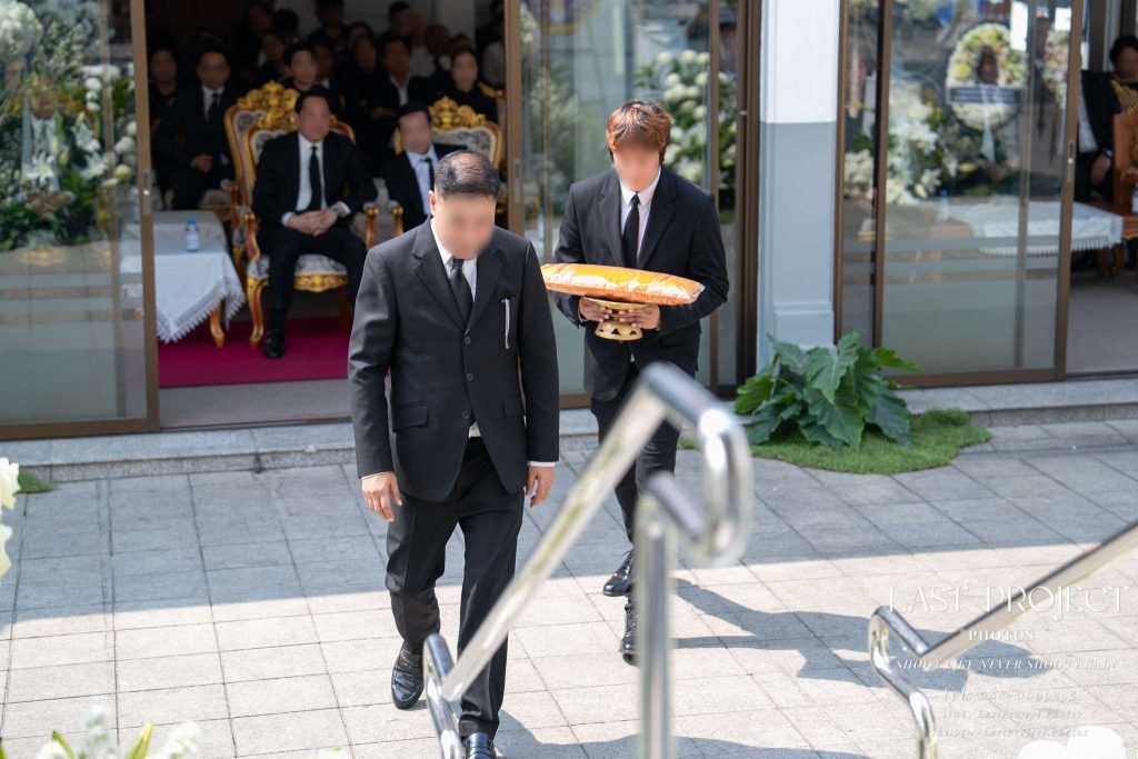 ภาพถ่ายพิธีพระราชทานเพลิงศพและฌาปนกิจ แขกผู้มีเกียรติถือพานผ้าไตรเดินขึ้นบันไดเมรุวัดไทย A solemn Thai Buddhist cremation ceremony at a temple showing a guest carrying monk robes on a phan pedestal tray up the crematorium steps.