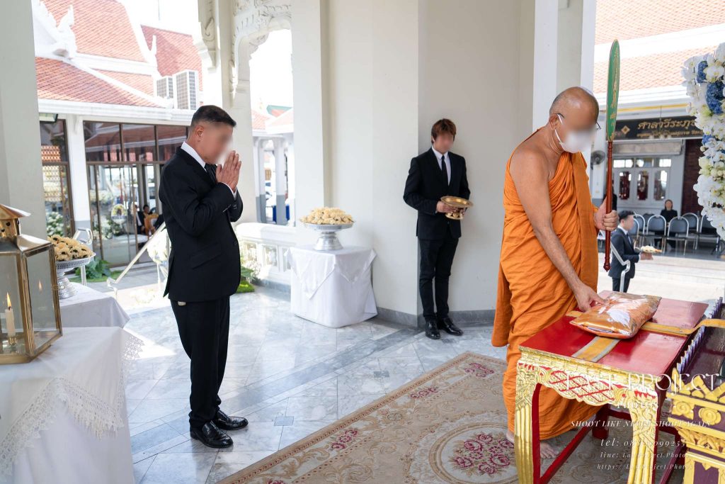 ภาพประธานในชุดสูทสีดำยืนพนมมือสำรวม ขณะพระสงฆ์ประกอบพิธีพิจารณาผ้าไตรบังสุกุลบนแท่นพิธีในงานฌาปนกิจ ณ ศาลาหน้าเมรุวัดไทย A host in a black suit standing with hands clasped in prayer as a Buddhist monk contemplates the Bangsukula robes on the ceremonial table during a cremation ritual at a Thai temple crematorium pavilion.
