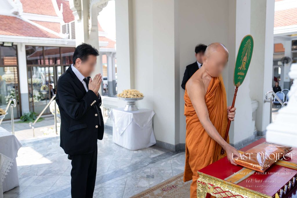 พระสงฆ์กำลังพิจารณาผ้าไตรบังสุกุลและแขกยืนประนมมือในพิธีฌาปนกิจศพ วัดเทพศิรินทร์ A Buddhist monk performing the Bangsukula ritual over a funeral robe with a guest praying respectfully at Wat Thepsirin