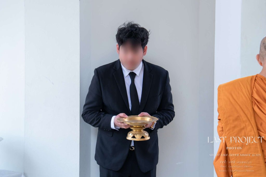 ภาพเจ้าภาพถือพานทองเตรียมถวายผ้าไตรบังสุกุลในพิธีฌาปนกิจศพ A host in a black suit holding a golden pedestal tray during a Buddhist funeral cremation ceremony