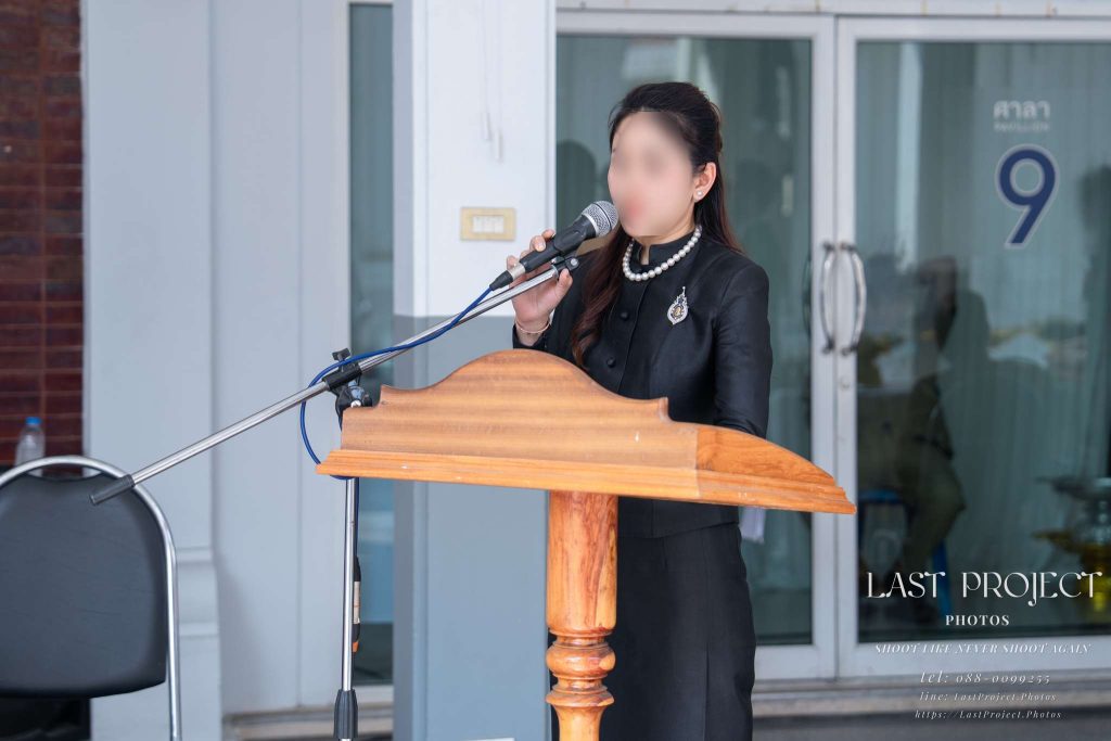 พิธีกรหรือญาติผู้วายชนม์กำลังกล่าวคำไว้อาลัยและประวัติผู้วายชนม์ที่โพเดียมในงานฌาปนกิจ A family member delivering a eulogy and biography at the podium during a Thai Buddhist funeral ceremony