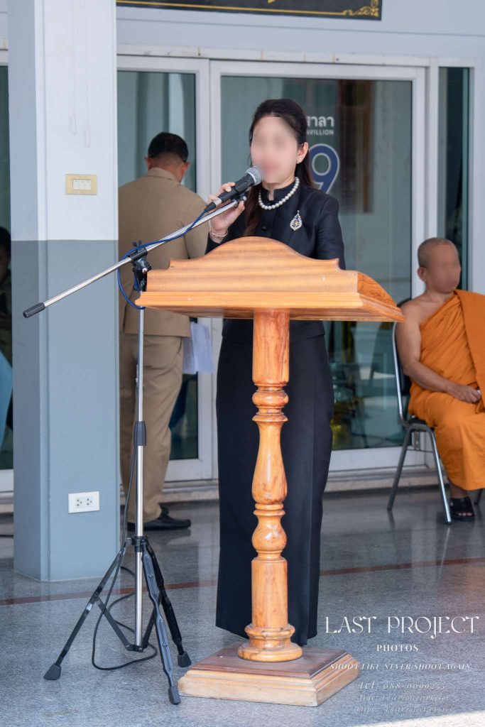 พิธีกรหญิงสวมชุดไทยจิตรลดาสีดำกล่าวคำไว้อาลัยและอ่านประวัติผู้วายชนม์หน้าโพเดียมในพิธีฌาปนกิจ A woman in a formal black Thai dress reading a eulogy at the wooden podium during a Buddhist cremation ceremony