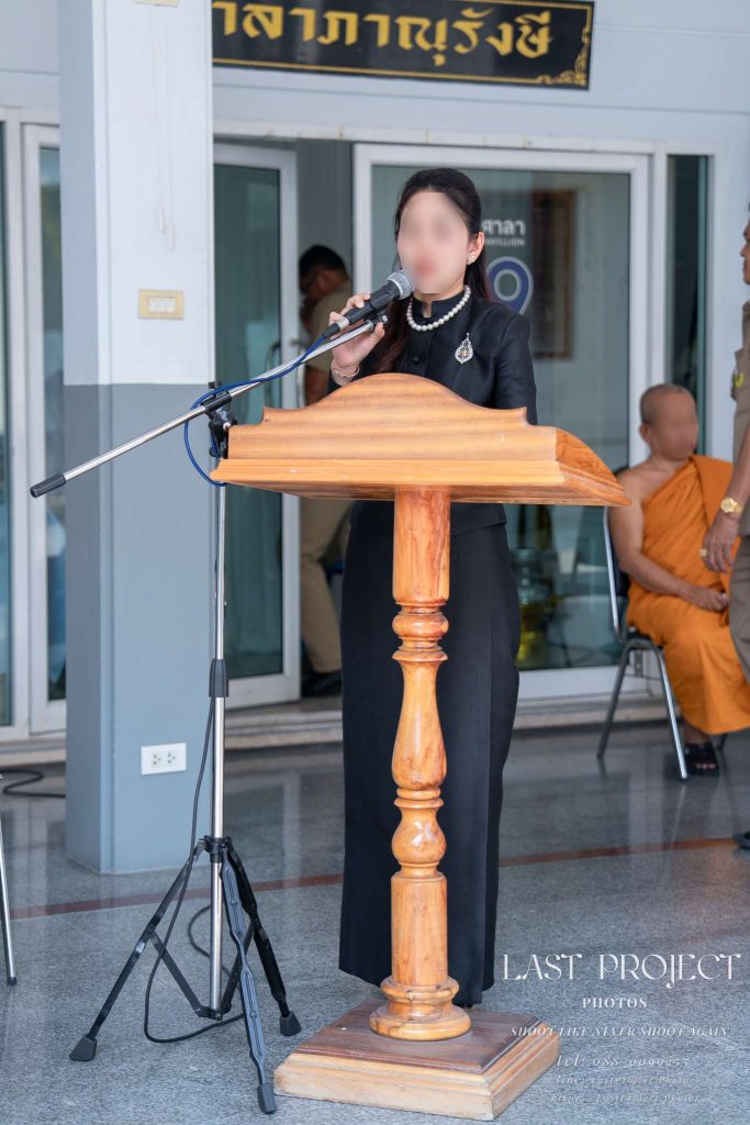 ภาพผู้หญิงสวมชุดจิตรลดาสีดำยืนพูดใส่ไมโครโฟนที่โพเดียมไม้ในงานพิธีศพ หน้าศาลาภาณุรังษี วัดเทพศิรินทร์ กรุงเทพมหานคร ด้านหลังมีพระสงฆ์นั่งอยู่บนเก้าอี้ A woman in black formal attire speaking at a wooden podium with a microphone during a funeral ceremony at Wat Debsirin, Bangkok, in front of the Bhanurangsi Pavilion with a Buddhist monk seated in the background.