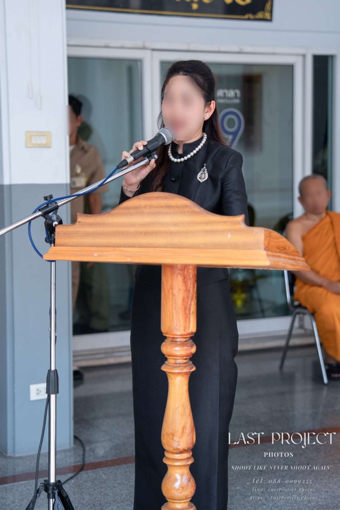 ภาพหญิงสาวในชุดดำจิตรลดาประดับเข็มกลัดไว้อาลัยกำลังกล่าวประวัติและคำไว้อาลัยแด่ผู้วายชนม์ที่โพเดียมไม้หน้าไมโครโฟนในพิธีฌาปนกิจ ณ วัดเทพศิรินทราวาสราชวรวิหาร โดยมีพระสงฆ์นั่งอยู่ด้านหลังและมีไมโครโฟนตั้งอยู่ด้านหน้า A woman in formal black attire reading a eulogy at a wooden podium during a Thai cremation ceremony at Wat Debsirindrawas Ratchaworawiharn, Bangkok, with a monk seated in the background.