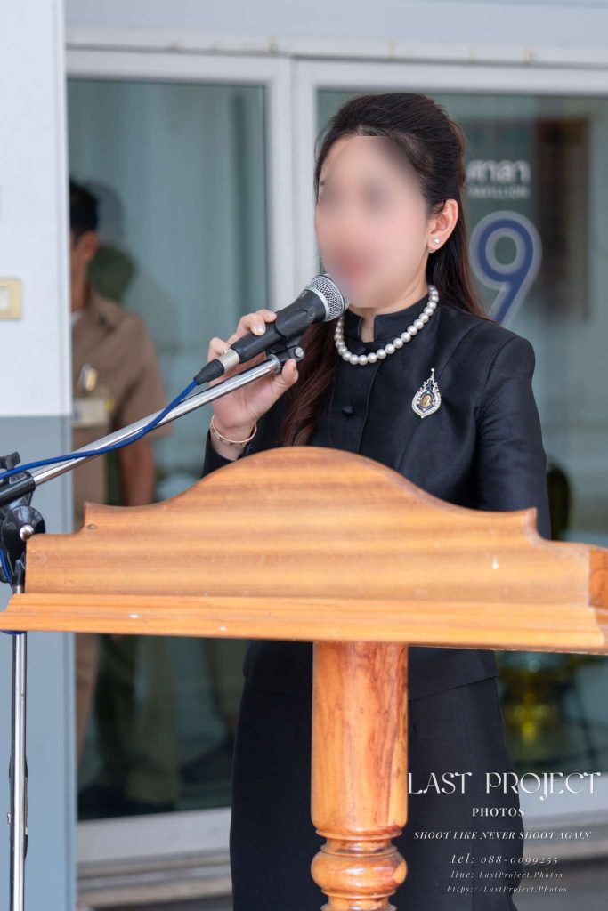 หญิงไทยในชุดไว้ทุกข์สีดำกำลังกล่าวคำไว้อาลัยและอ่านประวัติผู้วายชนม์ที่โพเดียมในพิธีฌาปนกิจศพ A Thai woman in traditional black mourning attire delivering a eulogy and reading the biography at the podium during a Buddhist cremation ceremony at a temple