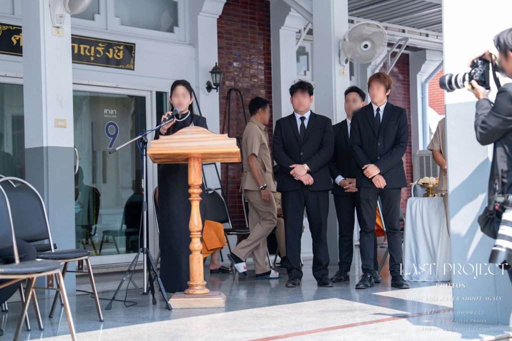 ภาพถ่ายพิธีฌาปนกิจศพขณะเจ้าภาพกล่าวชีวประวัติผู้วายชนม์หน้าศาลาวัด Buddhist cremation ceremony photograph capturing the host reading a memorial eulogy at the temple pavilion