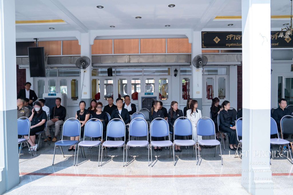 แขกผู้มีเกียรติและญาติมิตรนั่งร่วมพิธีฌาปนกิจศพอย่างสงบขณะรับฟังชีวประวัติผู้วายชนม์ ณ ศาลาวัด Guests and relatives seated quietly during a Thai Buddhist cremation ceremony at a temple pavilion