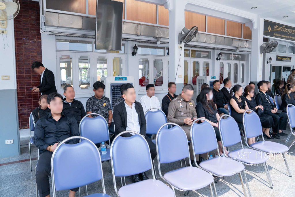 บรรยากาศผู้ร่วมงานสวมชุดดำและเครื่องแบบข้าราชการนั่งเก้าอี้พลาสติกสีน้ำเงินในศาลาวัดเพื่อรับฟังพิธีฌาปนกิจและประวัติผู้วายชนม์ในงานศพแบบไทย Attendees in black mourning attire and official uniforms sitting on blue chairs inside a Thai temple pavilion during a cremation funeral ceremony listening to the eulogy