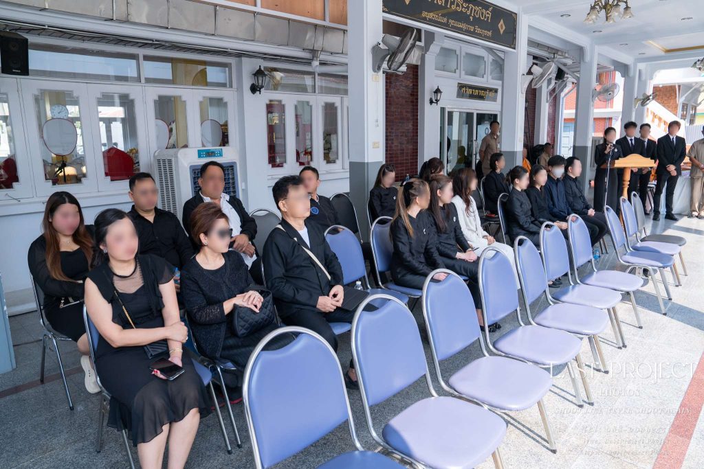 บรรยากาศแขกผู้ร่วมงานแต่งกายด้วยชุดไว้อาลัยสีดำนั่งบนเก้าอี้เพื่อฟังการอ่านประวัติผู้วายชนม์ในพิธีฌาปนกิจ ณ วัดเทพศิรินทร์ Guests dressed in black mourning attire seated on chairs listening to the eulogy during a Thai Buddhist cremation ceremony at Debsirindrawas Temple.