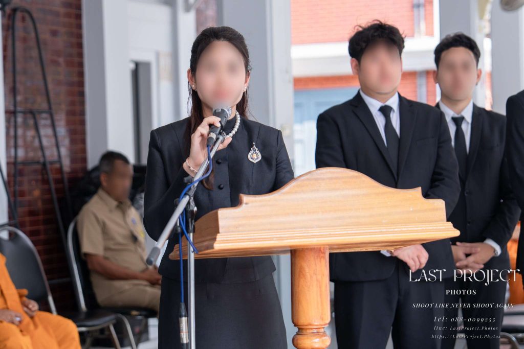 ภาพถ่ายเจ้าภาพหญิงยืนอ่านประวัติและคำไว้อาลัยผู้วายชนม์ที่โพเดียมด้านหน้าศาลาบำเพ็ญกุศลในพิธีฌาปนกิจศพแบบไทย Female host delivering a eulogy at the podium during a Thai Buddhist cremation ceremony in front of the funeral pavilion