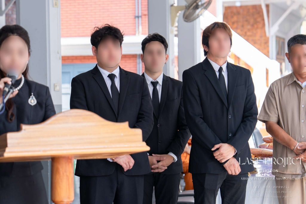 ภาพญาติและแขกผู้ร่วมงานยืนไว้อาลัยด้านหลังพิธีกรขณะอ่านประวัติผู้วายชนม์ในพิธีฌาปนกิจศพ ณ วัดไทย Close relatives and guests wearing black mourning attire standing solemnly behind a speaker reading a eulogy during a Thai Buddhist cremation ceremony at a temple.
