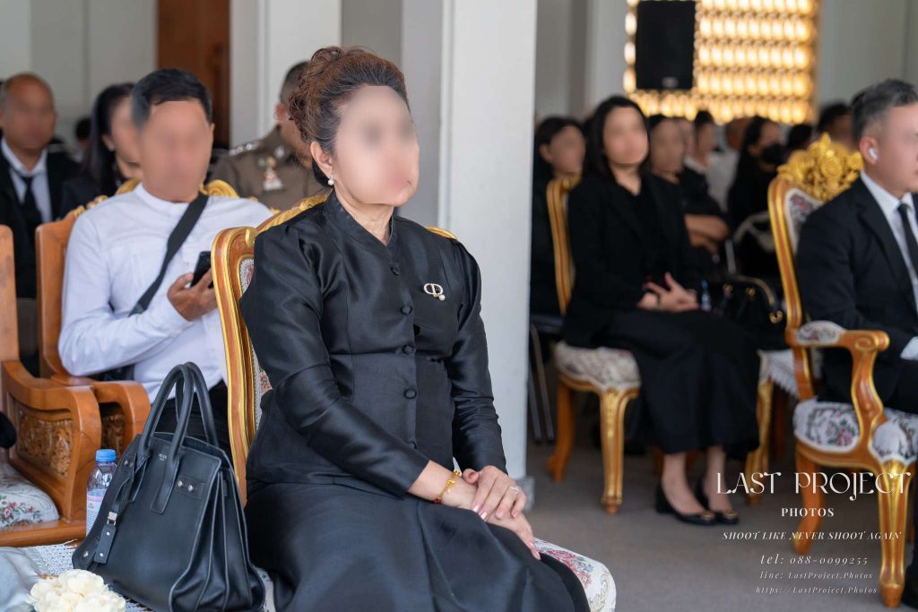 ภาพถ่ายแขกสวมชุดดำไว้อาลัยกำลังนั่งฟังการอ่านชีวประวัติในพิธีฌาปนกิจศพ Female guest in formal black mourning attire attentively listening to a eulogy during a Thai Buddhist cremation ceremony